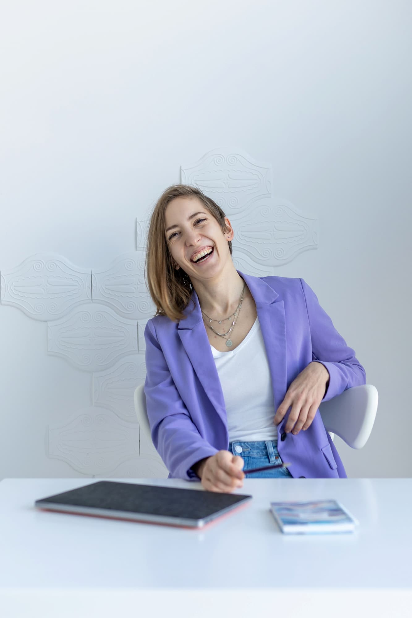 Mina is sitting at a table wearing a light blue blouse with a white shirt. They head is tilted, smiling into the camera. They have shoulder-length hair. In their hands, they are holding a pen, and a laptop and note book sit in front of them.